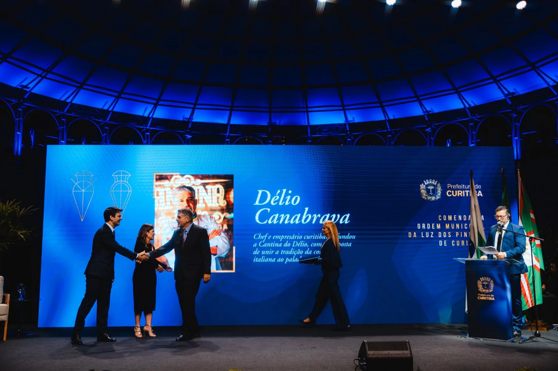 Délio Canabrava no palco da cerimônia da Comenda da Ordem Municipal da Luz dos Pinhais, com seu nome no telão